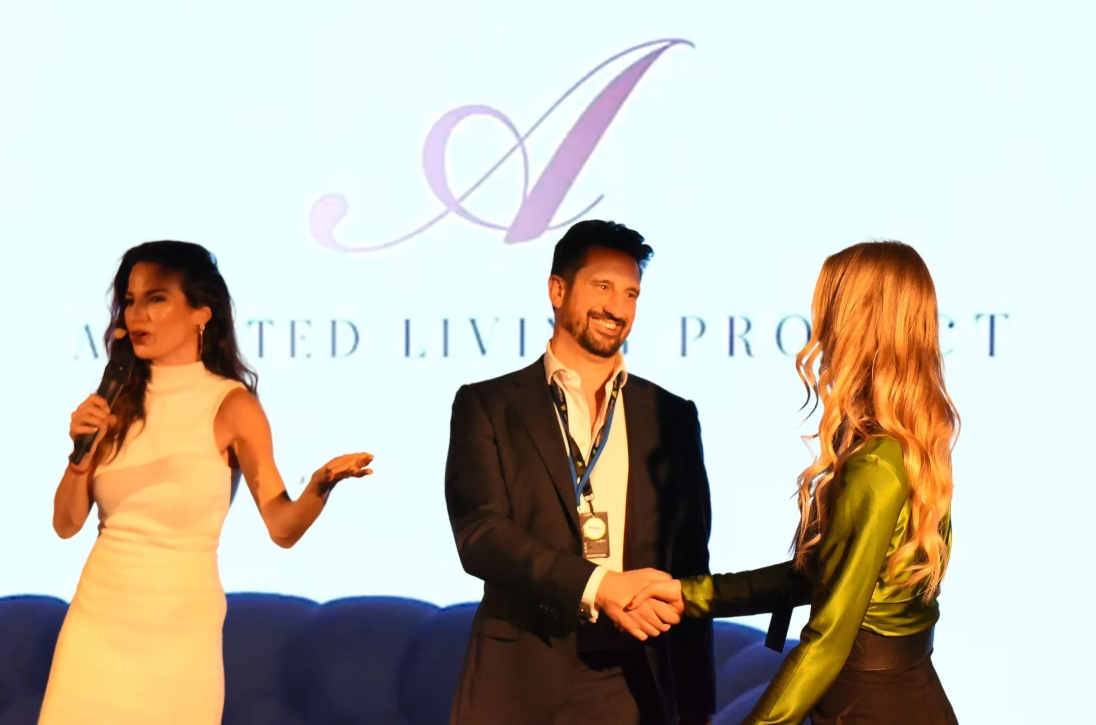 Thomas Williams shaking hands on stage as he accepts the Best Pitch award, the Assisted Living Project name projected on the screen behind him